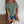 Vrouw in groene geperforeerde blouse en denim shorts, met bruine schoudertas en koffiehouder, tegen witte bakstenen muur. Casual zomeroutfit.