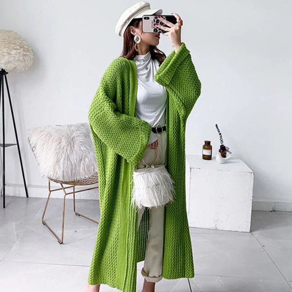 Vrouw in groene gebreide oversized cardigan, witte coltrui en beige broek, poseert met smartphone. Modieuze outfit, trendy herfstkleding.