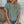 Vrouw draagt groene geperforeerde blouse met korte mouwen en denim shorts, zittend tegen een witte bakstenen muur. Casual zomeroutfit, modetrend 2023.