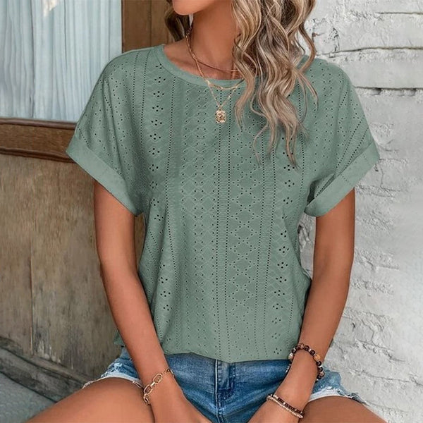 Vrouw draagt groene geperforeerde blouse met korte mouwen en denim shorts, zittend tegen een witte bakstenen muur. Casual zomeroutfit, modetrend 2023.