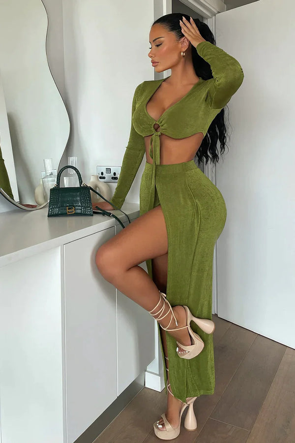Vrouw in groene jurk met hoge split, poseert naast witte kast. Stijlvolle mode, trendy outfit, hoge hakken, moderne interieurstijl.