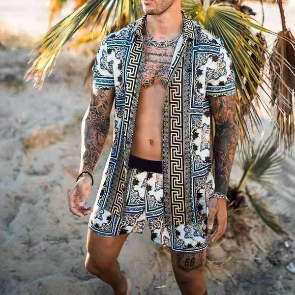 Man in kleurrijke, gedessineerde zomeroutfit met korte mouwen en shorts, staand op een zandstrand met palmbomen. Perfect voor strandmode en zomerse stijl.