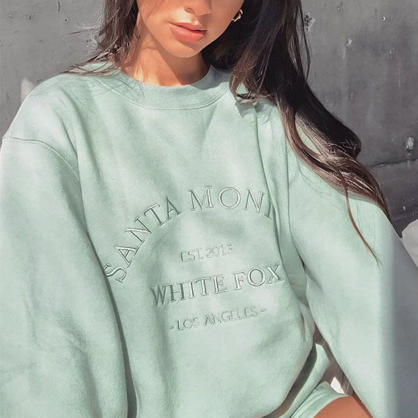 Vrouw in mintgroene oversized trui met opdruk "Santa Monica White Fox Los Angeles", casual mode, trendy streetwear, zonnige achtergrond.
