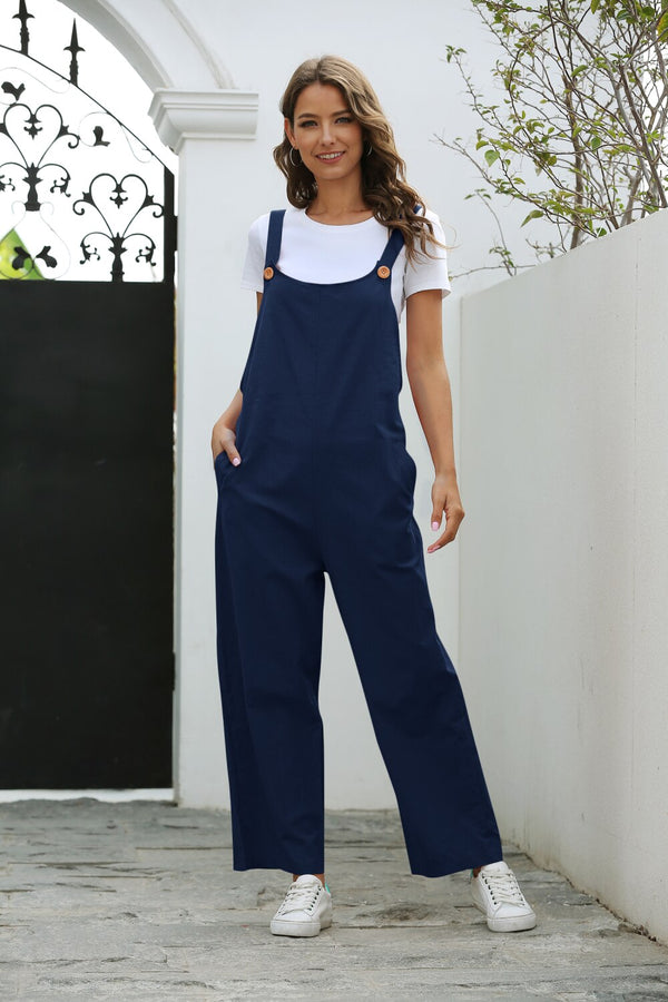 Vrouw in blauwe tuinbroek en witte T-shirt poseert buiten bij witte muur en smeedijzeren poort, casual mode, comfortabele dameskleding.