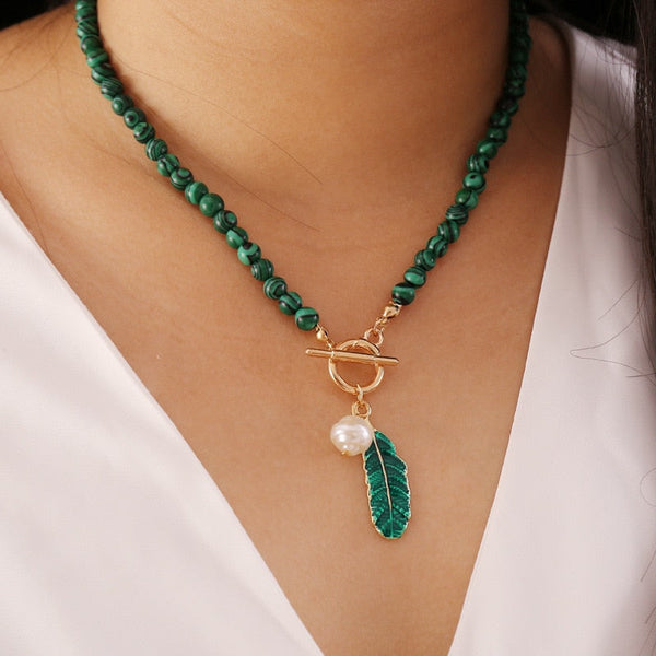 Groene kralenketting met gouden sluiting, parelhanger en groene bladbedel, gedragen op witte blouse. Elegante sieraden voor dames.