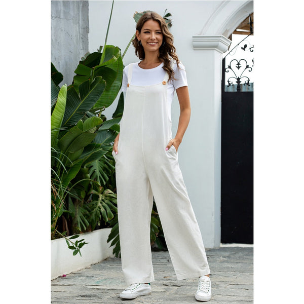 Vrouw in witte jumpsuit en sneakers poseert buiten bij groene planten en witte muur. Casual mode, zomerse outfit, comfortabele kledingstijl.
