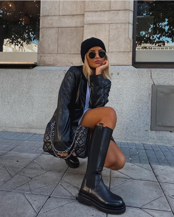 Vrouw in zwarte leren jas en laarzen, zittend op stoep. Draagt een zwarte muts en zonnebril. Modieuze herfstoutfit, stedelijke stijl.