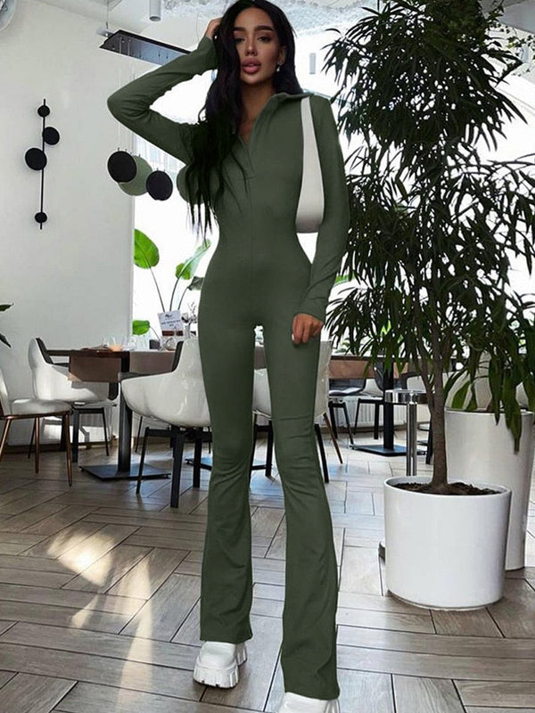Vrouw in groene jumpsuit met witte tas, staand in moderne caféomgeving met planten en witte stoelen. Trendy mode, casual outfit, stedelijke stijl.