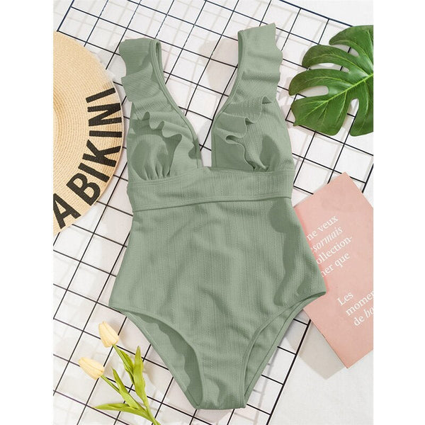 Groen badpak met ruches op een geruite achtergrond, naast een strohoed met "BAIKINI" en een roze boek. Perfect voor strandmode en zomertrends.