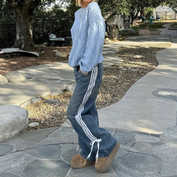 Vrouw in blauwe trui en wijde spijkerbroek met zijstrepen en veterdetails, buiten op een stenen pad. Trendy Y2K mode, casual en comfortabel.