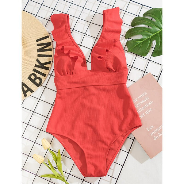Rood badpak met ruches, zomerse strandstijl, naast een strohoed en groene bladeren, op een geruite achtergrond. Perfect voor strandmode.