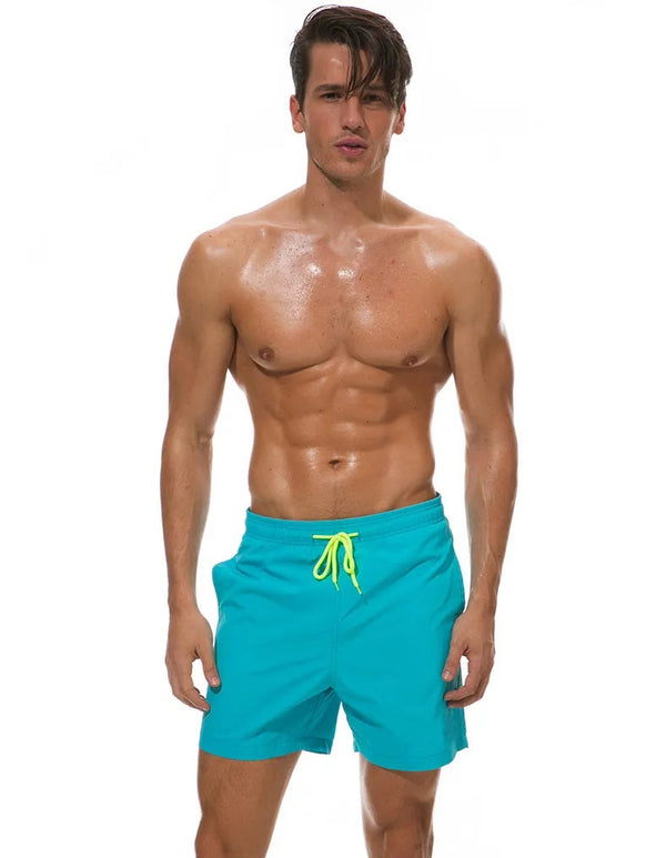 Man in turquoise zwembroek met gele koord, gespierd en bezweet, poseert voor witte achtergrond. Perfect voor strandmode en fitness inspiratie.