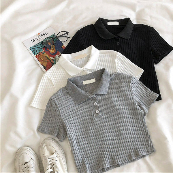 Drie geribbelde poloshirts in zwart, wit en grijs op een bed met een Matisse-boek en bril. Modieuze dameskleding, casual stijl.