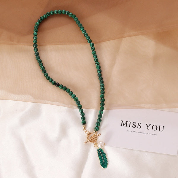 Groene kralenketting met gouden sluiting en bladbedel, naast een kaart met de tekst "MISS YOU". Perfecte sieradenaccessoire voor dames.