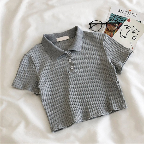 Grijs geribd poloshirt met korte mouwen op een witte achtergrond, naast een bril en een Matisse-boek. Modieuze dameskleding, casual stijl.
