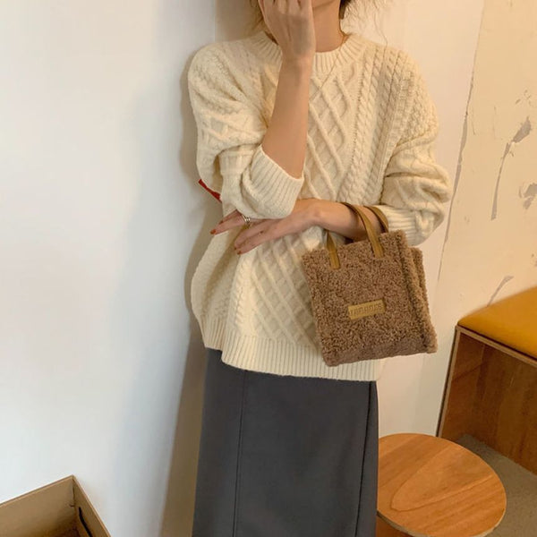 Vrouw in crèmekleurige kabeltrui met bruine teddy handtas, leunend tegen muur. Modieuze herfstoutfit, casual stijl, trendy accessoires.