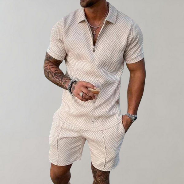Man in beige zomerset met geometrisch patroon, korte mouwen en korte broek, drinkt een drankje. Stijlvolle herenkleding voor de zomer.