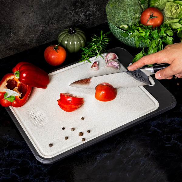 Hand snijdt tomaat op snijplank met mes, omringd door paprika, knoflook, tomaten, broccoli en kruiden. Keuken, koken, verse groenten.