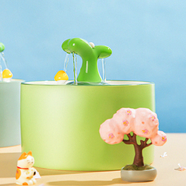 Groene kattenwaterfontein met speelse gele eend, omringd door decoratieve items zoals een boompje en een kattenbeeldje, tegen een blauwe achtergrond.