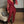 Vrouw in elegante rode jumpsuit met doorschijnende mouwen, staand naast een beige stoel. Perfect voor avondkleding of feestelijke gelegenheden.