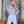 Vrouw in elegante lichtblauwe jumpsuit met veren en glinsterende details, poseert in moderne interieursetting met planten en houten elementen.