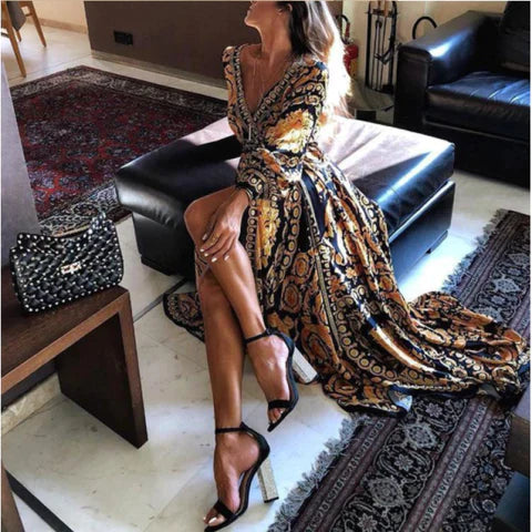 Vrouw in elegante geel-zwarte jurk met barokprint, zittend op zwarte bank. Stijlvolle outfit, hoge hakken, luxe interieur, modetrend.
