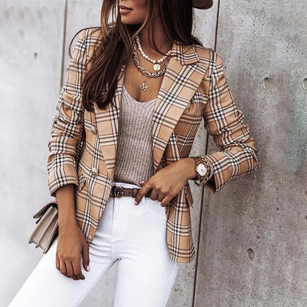 Vrouw in modieuze kaki geruite blazer, beige top en witte broek, leunt tegen betonnen muur. Stijlvolle herfstoutfit, trendy damesmode.