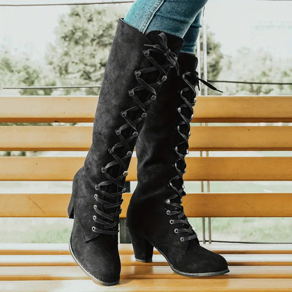 Zwarte suède laarzen met hoge hak en veters, gedragen door een persoon in blauwe jeans, op een houten bank. Trendy damesmode, herfststijl.