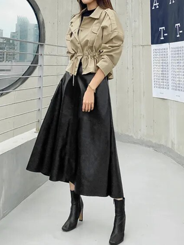 Vrouw in beige jas en zwarte midi-rok, staand op balkon met stadzicht. Modieuze herfstoutfit, trendy dameskleding, stedelijke stijl.