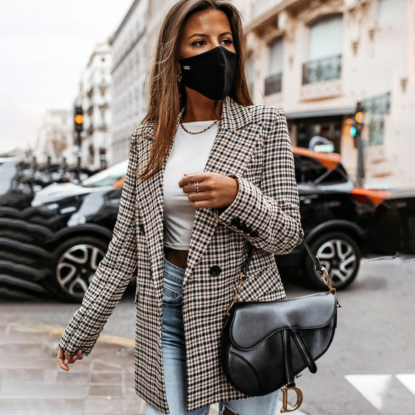 Vrouw met zwart mondkapje, geruite blazer en zwarte schoudertas loopt op straat. Mode, herfstkleding, stedelijke stijl.