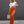 Vrouw in trendy outfit met witte gebreide crop top en oranje flared broek, draagt zonnebril en beige handtas, poseert voor grijze achtergrond.