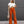 Vrouw in trendy outfit met witte gebreide crop top en oranje flared broek, draagt zonnebril en beige tas, poseert voor grijze achtergrond.