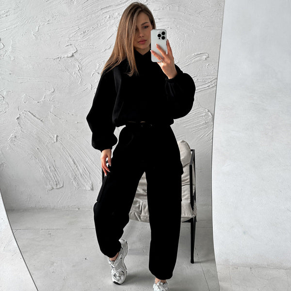 Vrouw in zwarte oversized trui en broek poseert voor spiegel, houdt smartphone vast. Modieuze casual outfit, trendy sneakers, minimalistisch interieur.