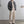 Man in casual outfit met grijze geribbelde trui, crèmekleurige broek en geruite schoenen, staand in minimalistische ruimte met kopjes op tafel.