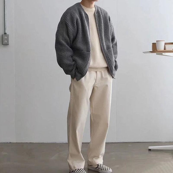 Man in casual outfit met grijze geribbelde trui, crèmekleurige broek en geruite schoenen, staand in minimalistische ruimte met kopjes op tafel.
