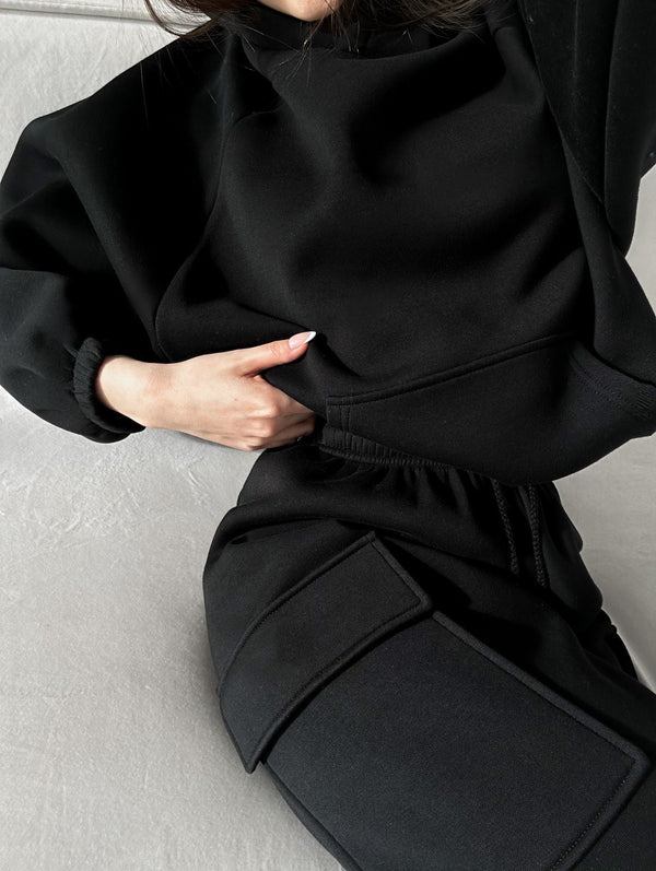 Vrouw in zwarte oversized hoodie en joggingbroek, ontspannen pose op grijze achtergrond. Comfortabele loungewear, modieuze casual kledingstijl.