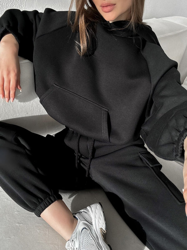 Vrouw in zwarte oversized hoodie en joggingbroek, zittend op witte bank. Casual sportieve mode, comfortabele loungewear, grijze sneakers.