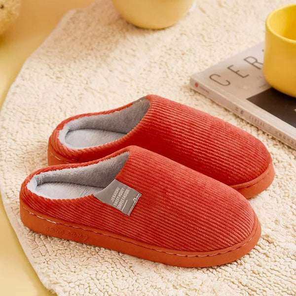 Oranje ribfluwelen pantoffels op beige tapijt, naast gele mok en tijdschrift. Comfortabele huisschoenen, perfect voor herfst en winter.
