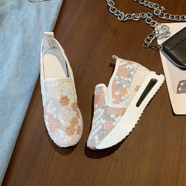 Roze en witte damessneakers met bloemenpatroon op houten vloer, naast een zilveren ketting en blauwe jeans. Comfortabele en modieuze schoenen.