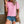 Vrouw in roze geperforeerd T-shirt en denim shorts, met bruine schoudertas. Casual zomeroutfit, trendy damesmode, buitenmuur achtergrond.