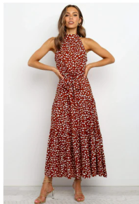 Vrouw in lange, mouwloze rode jurk met witte bloemenprint, halternek en riem, poseert tegen witte achtergrond. Trendy damesmode, zomerjurk.