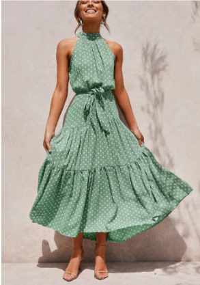Vrouw in groene polkadot zomerjurk met halternek en strikceintuur, poseert tegen een neutrale achtergrond. Perfecte outfit voor zomermode.