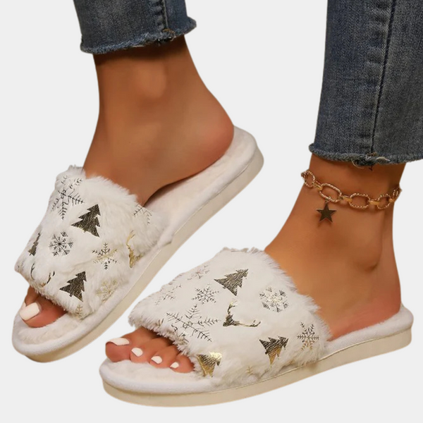 Witte pluche slippers met kerstboom- en sneeuwvlokmotief, gedragen door een persoon met jeans en een stervormige enkelband. Perfect voor wintermode.