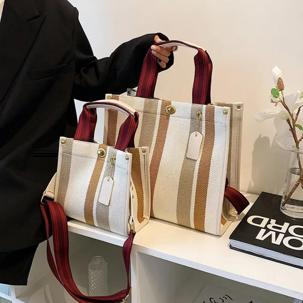 Twee gestreepte canvas handtassen met rode handvatten op een witte tafel, naast een vaas met bloemen. Stijlvolle damesaccessoires, modetrend.
