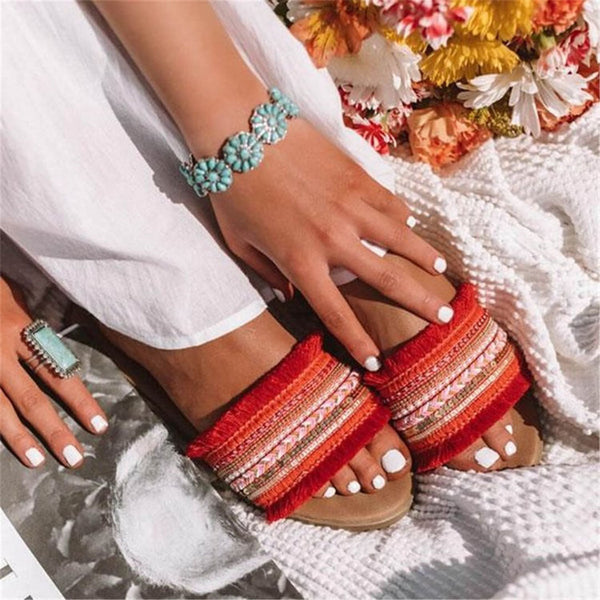 Vrouw met rode sandalen en turquoise sieraden op witte deken, omringd door bloemen. Zomerschoenen, boho-stijl, kleurrijke accessoires.