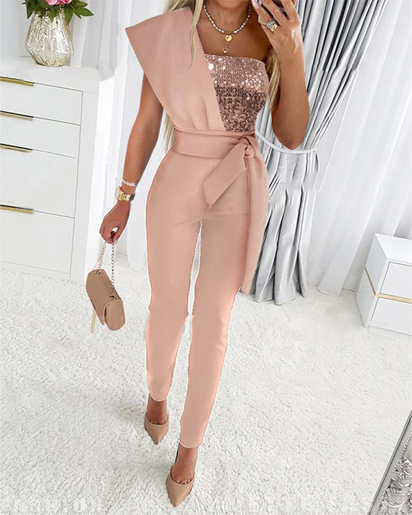 Vrouw in roze jumpsuit met pailletten top, asymmetrisch ontwerp, strikceintuur, hoge hakken en handtas, staand in moderne slaapkamer.