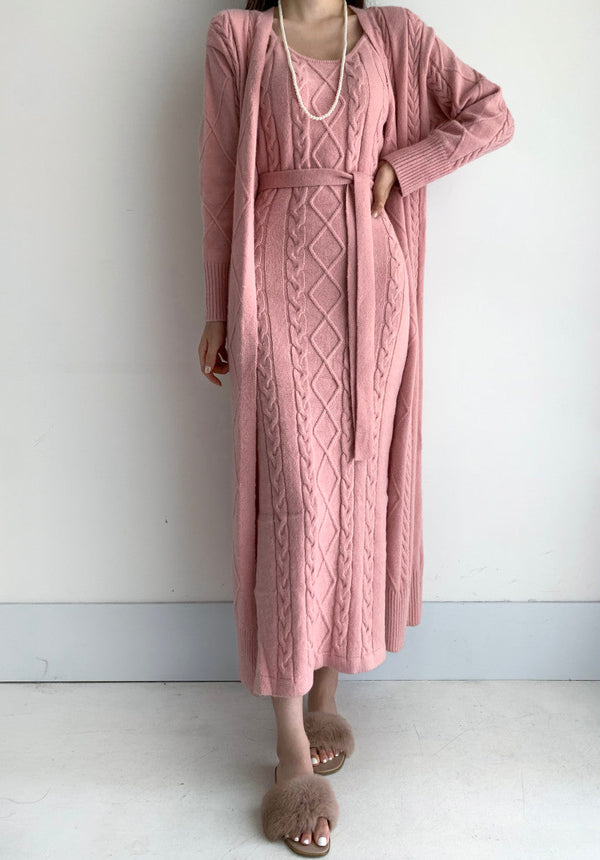 Roze gebreide jurk met kabelpatroon, lange mouwen en ceintuur, gecombineerd met bijpassend vest en parelketting. Elegante damesmode outfit.