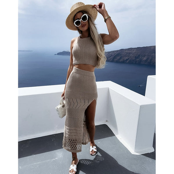 Vrouw in beige gehaakte crop top en rok met split, draagt zonnebril en strohoed, poseert op wit terras met uitzicht op zee en kliffen. Zomermode.