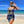 Vrouw in blauwe zomerjurk met gouden kettingprint op tropisch strand, zonnebril, blauwe lucht, zee. Perfecte strandoutfit, modetrend 2023.