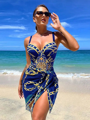 Vrouw in blauwe zomerjurk met gouden kettingprint op tropisch strand, zonnebril, blauwe lucht, zee. Perfecte strandoutfit, modetrend 2023.